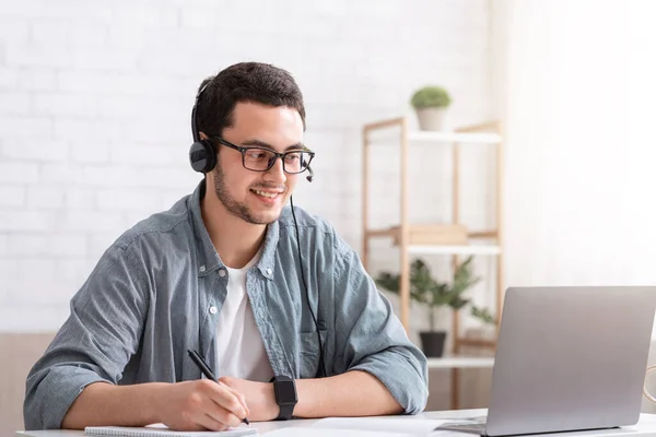 Ajuda ao cliente remotamente. Consultor sorridente cara em óculos e fone de ouvido olha para laptop e faz anotações no notebook — Fotografia de Stock