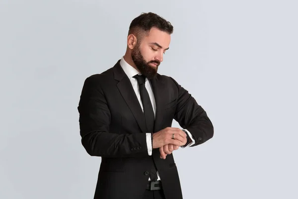 Handsome young businessman in black suit checking time on his watch on grey background — Stock Photo, Image