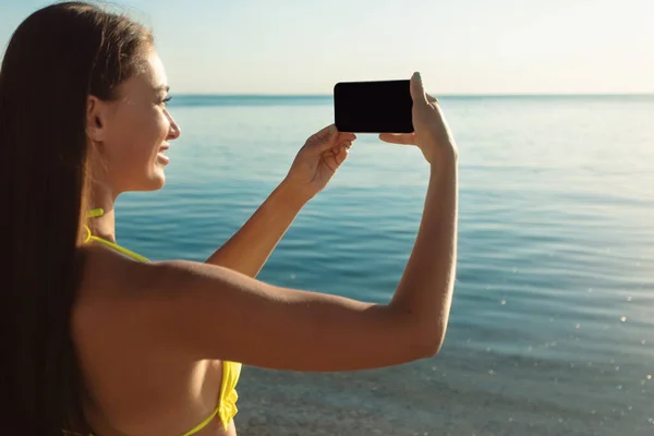 Κορίτσι σε μαγιό κρατώντας το τηλέφωνο λήψη φωτογραφιών της θάλασσας σε εξωτερικούς χώρους — Φωτογραφία Αρχείου