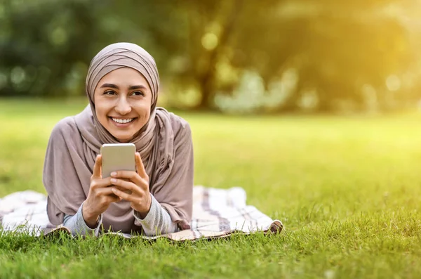 Mujer musulmana riéndose usando su teléfono inteligente, descansando en el parque —  Fotos de Stock