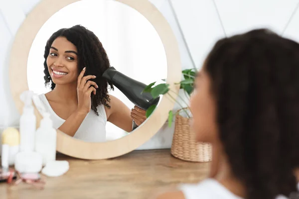美丽的黑人妇女在镜子前与吹风机一起晒干卷发 — 图库照片