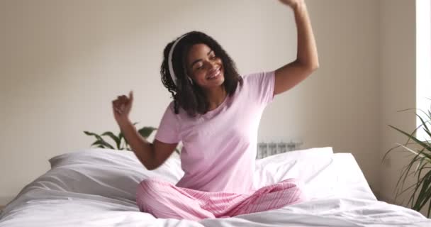 Energisk svart kvinna dansar i sängen, lyssnar på musik — Stockvideo