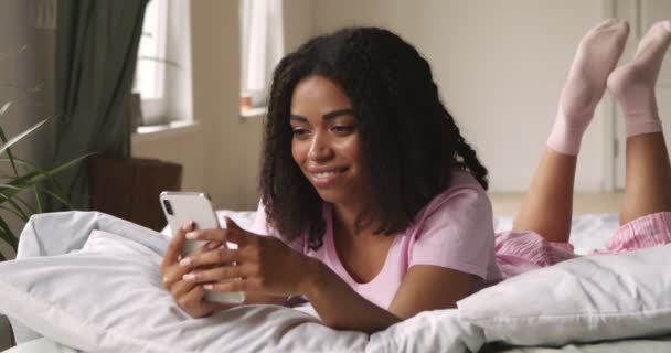 Svart flicka chattar med vänner på mobilen, liggande i sängen — Stockvideo
