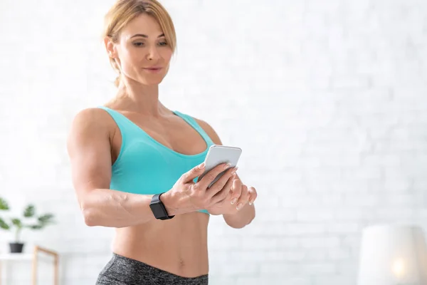 Entrenamiento en línea. Mujer con reloj inteligente comprueba teléfono inteligente en la sala de estar —  Fotos de Stock