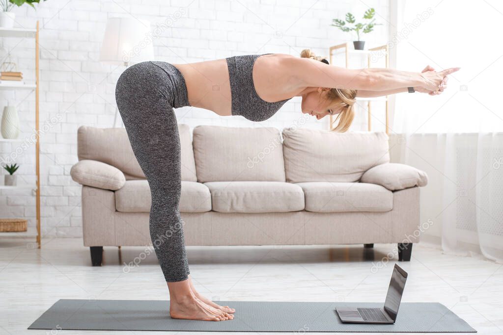 Fitness online at home during epidemic. Woman makes tilt, standing on mat with laptop