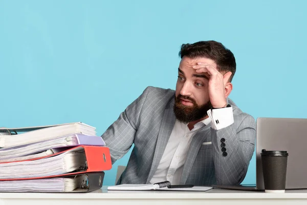 Exhausted millennial businessman overwhelmed with paperwork at desk with laptop on blue background — Stock Photo, Image