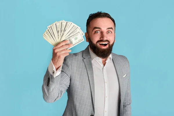 Alegre hombre de negocios barbudo en traje con abanico de billetes de dólar sobre fondo azul —  Fotos de Stock