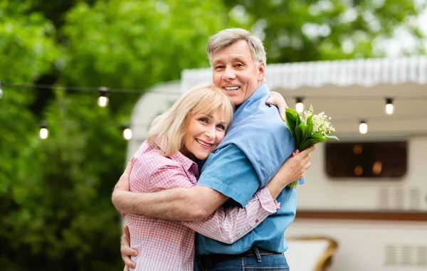 Szczęśliwy Dojrzałe para z bukiet z pola kwiaty przytulanie w pobliżu przyczepy na zewnątrz — Zdjęcie stockowe