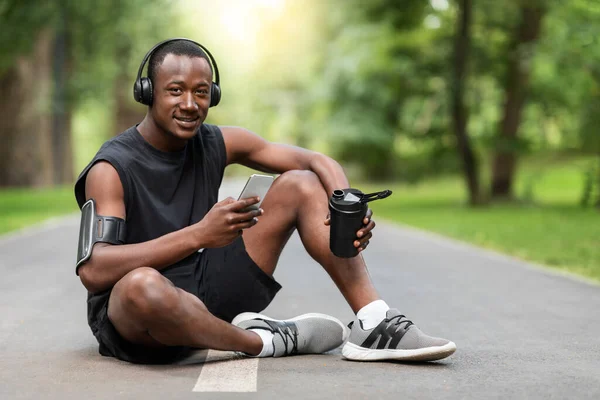 Deportista africano sentado en el camino del parque, bebiendo y usando smartphone —  Fotos de Stock