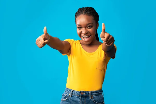 Радостная чернокожая девушка с большими пальцами, глядя на камеру, синий фон — стоковое фото