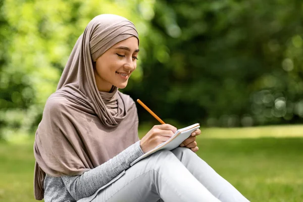 Chica en hijab escribiendo en el cuaderno, estudiando en el parque público — Foto de Stock