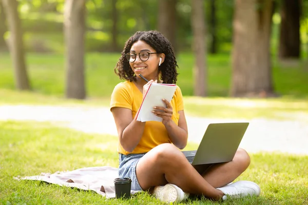 Éducation en ligne. Heureuse fille noire prenant des notes tout en regardant la leçon web sur ordinateur portable au parc — Photo