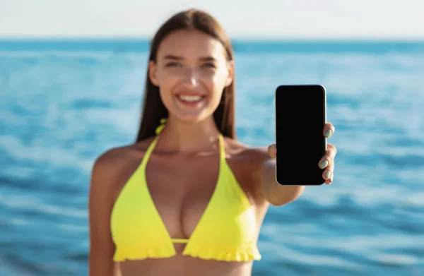 Chica en traje de baño mostrando la pantalla del teléfono inteligente de pie en la playa al aire libre — Foto de Stock