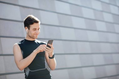 Müzikle modern çalışma. Telefonla gülümseyen genç adam, fitness takip cihazı ve kablosuz kulaklıklar antremana hazırlanıyor.