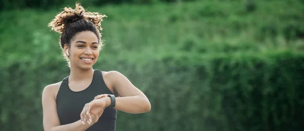 Ejercicio al aire libre. Sonriente chica afroamericana con auriculares inalámbricos comprueba rastreador de fitness —  Fotos de Stock