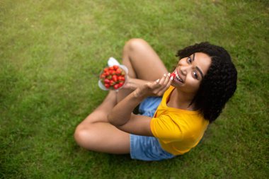 Sağlıklı beslenme. Yeşil çimenlikte lezzetli çilekler yiyen zinde siyah kız manzarasının üstünde, boşluğu kopyala