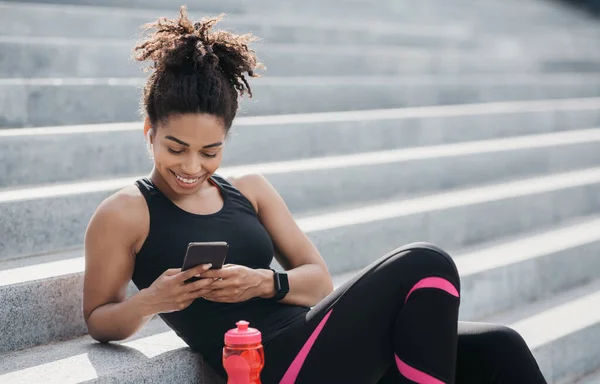 Chica afroamericana en ropa deportiva con rastreador de fitness se ve en el teléfono inteligente, cerca de la botella de agua —  Fotos de Stock