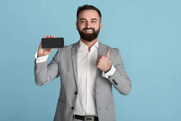 Glückliche Büroangestellte hält Smartphone mit leerem Bildschirm und zeigt Daumen nach oben auf blauem Hintergrund, Platz für Design — Stockfoto