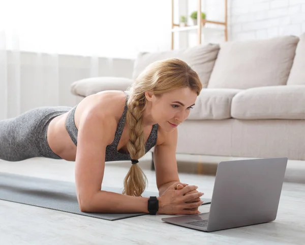 Frau mittleren Alters in Sportbekleidung mit Fitness-Tracker macht Planke auf Matte am Boden und blickt auf Laptop — Stockfoto