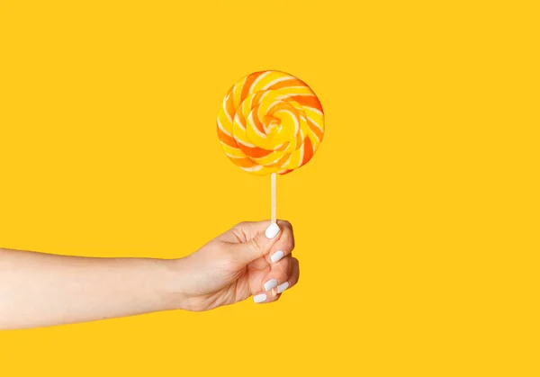Young woman holding swirl lollipop over orange background, close up of hand — Stock Photo, Image