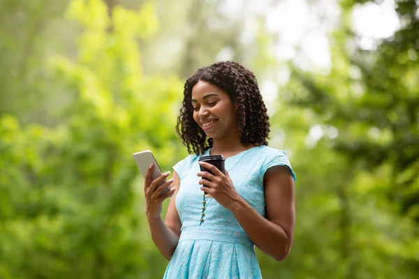 Mooie zwarte dame met afhaal koffie browsen sociale media op mobiele telefoon in het park — Stockfoto