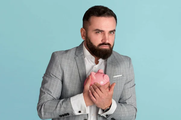 Ganancioso jovem empresário abraçando porquinho banco contra fundo azul — Fotografia de Stock