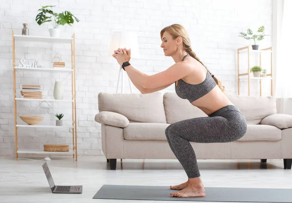 Mujer con rastreador de fitness crouches y relojes en el ordenador portátil en el interior de la sala de estar —  Fotos de Stock