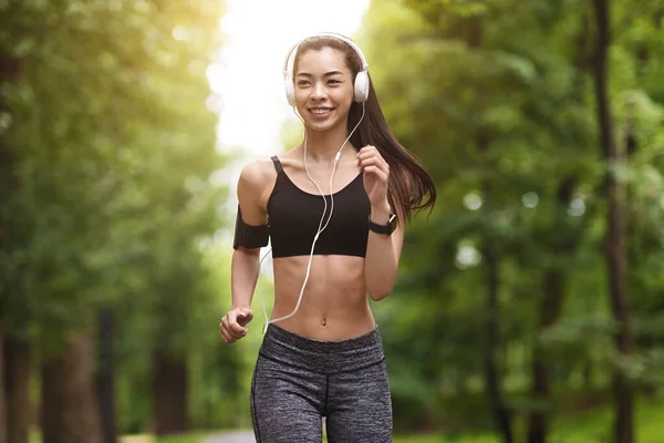 Morgenlauf. Lächelnde asiatische Mädchen mit Kopfhörern joggen im grünen Park — Stockfoto