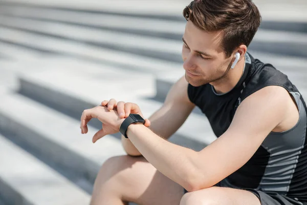 Aparatos modernos para correr. Atleta cansado con auriculares inalámbricos mira el rastreador de fitness y se sienta en los pasos —  Fotos de Stock