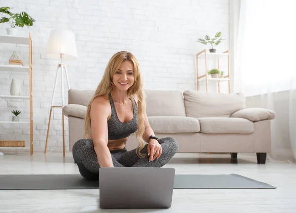 Uśmiechnięta kobieta w odzieży sportowej patrzy na laptopa, siedzi na podłodze na macie sportowej we wnętrzu salonu — Zdjęcie stockowe
