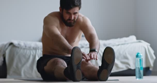 Молодой человек растягивает свое тело, сидит на полу дома — стоковое видео