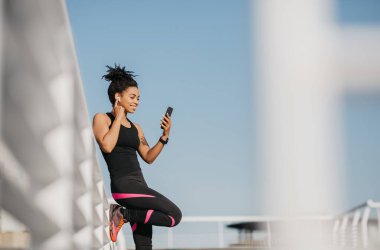 Koşu müziği. Spor giyim sektöründeki Afrikalı Amerikalı kız akıllı telefona kablosuz kulaklık ve saat takıyor.