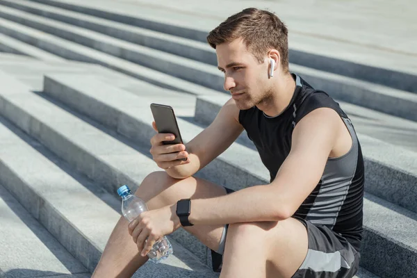 Atleta con cuffie wireless e fitness tracker tiene in mano bottiglia d'acqua e smartphone, si siede sulle scale — Foto Stock