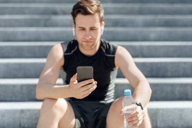 Spor kıyafetli, kulaklıklı, akıllı telefonlara bakıp su tutan, merdivenlerde oturan yorgun bir adam.