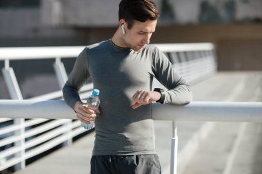 Kulaklık ve su şişeli kaslı adam spor saatine bakıyor.