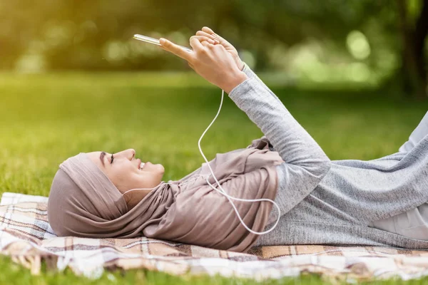 Neşeli Müslüman kız parkta dinleniyor, akıllı telefondan video izliyor. — Stok fotoğraf
