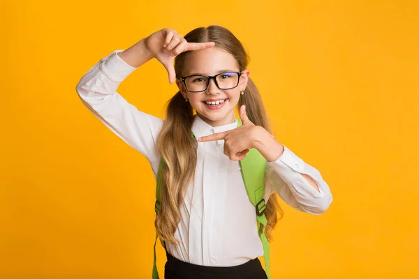 Vrolijk schoolmeisje op zoek naar camera door middel van vingers Frame, Studio schot — Stockfoto