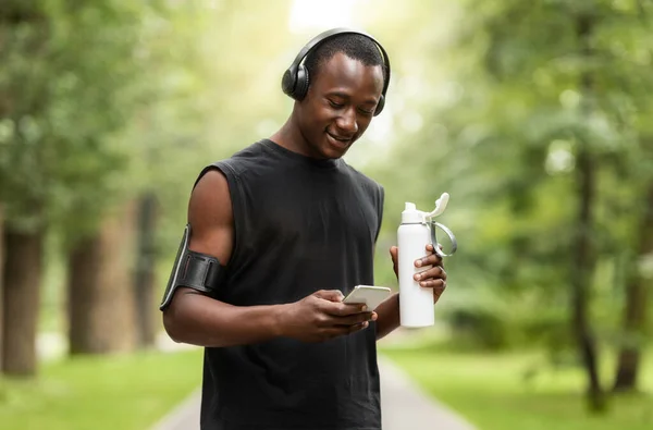 Chico negro feliz usando teléfono y agua potable, entrenamiento al aire libre —  Fotos de Stock