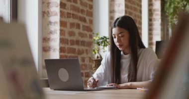 Asyalı öğrenci notlar alıyor, laptop ekranına bakıyor, profesörü takip ediyor.