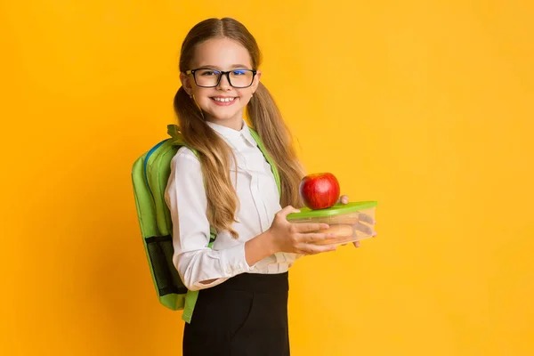 Školačka drží školní oběd box a červené jablko, žluté pozadí — Stock fotografie