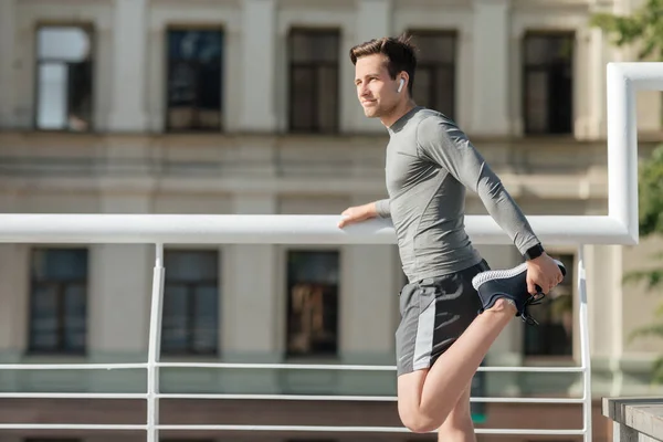 Young muscular man in sportswear and wireless headphones doing stretch for leg