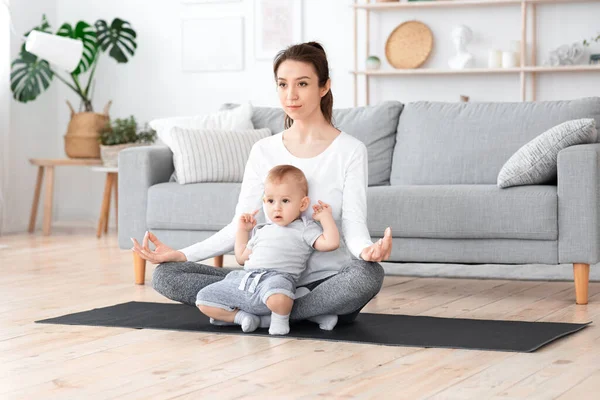 Uwolnienie stresu w macierzyństwie. Młoda mama medytuje w domu z dzieckiem na kolanach — Zdjęcie stockowe