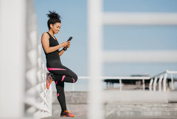 Chica afroamericana en uniforme deportivo se apoya en la pared con auriculares inalámbricos y mira el teléfono —  Fotos de Stock