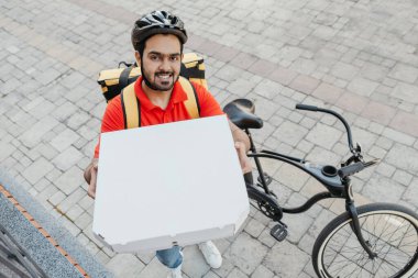 Sipariş teslimi. Kasklı, sakallı, gülümseyen adam, gözetleme deliğine pizza kutusu gösteriyor.