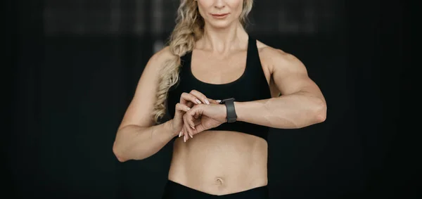 Resultado del entrenamiento. Musculosa mujer adulta en sujetador deportivo, mira el rastreador de fitness en el brazo sobre fondo oscuro —  Fotos de Stock