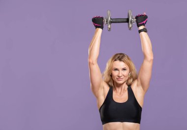 Dambılla egzersiz yap. Spor giyimli, kaslı, olgun bir kadın. Eldivenleri ve akıllı saati var. Dambılı başının üstünde tutuyor.