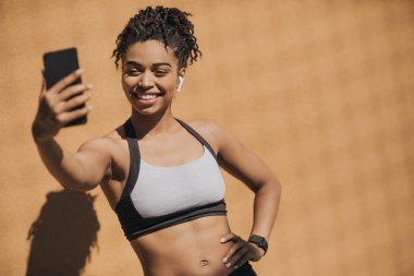 Kablosuz kulaklıkları ve fitness takip cihazıyla selfie çeken spor sutyenli Afrikalı Amerikalı genç bir kadın.