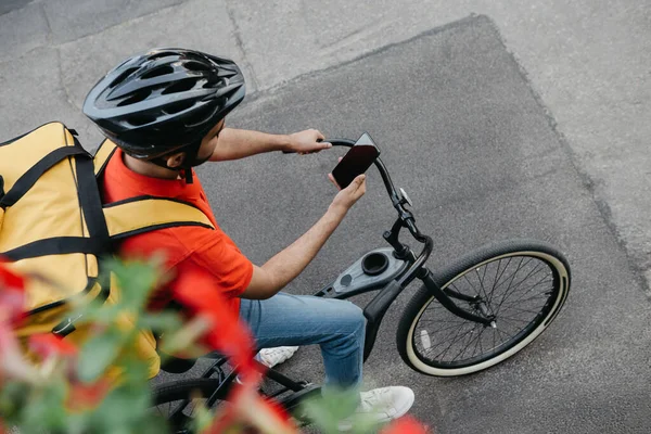 Cyklistický kurýr v ochranné přilbě s doručovacím batohem sedí na kole a dívá se na telefon a používá on-line mapu na ulici — Stock fotografie