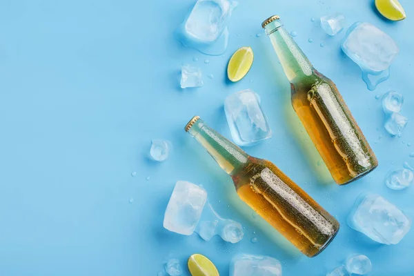 Две холодные бутылки светлого пива без этикетки со льдом и кусочками лайма на синем фоне — стоковое фото