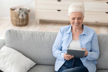 Evdeki kanepede dijital tablet bilgisayar kullanarak oturan çekici yaşlı kadın.